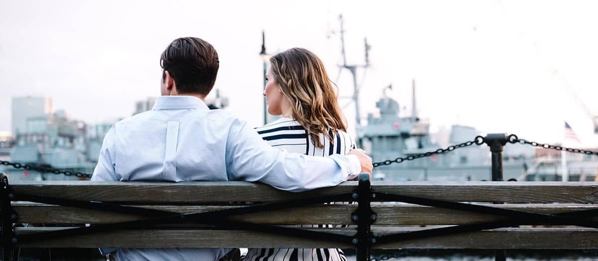 A man with his arm wrapped around a woman while sitting on a bench overlooking a body of water with a large ship docked.