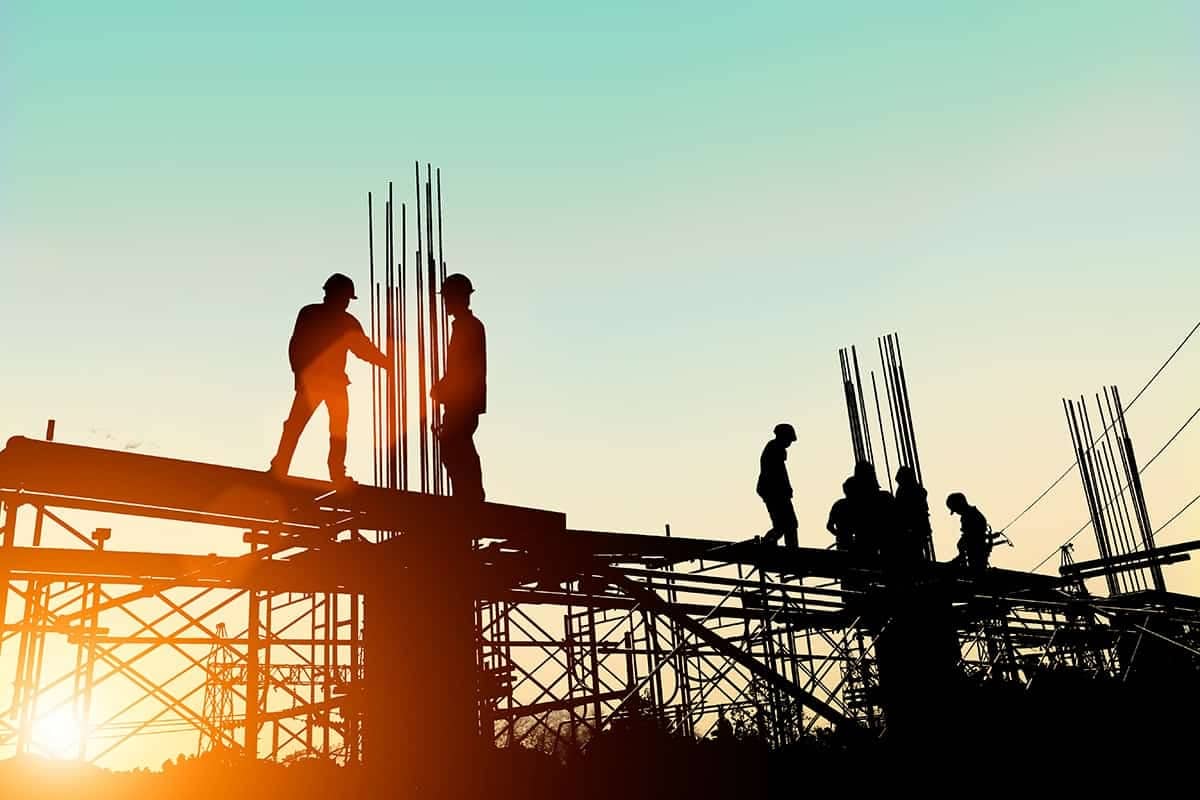 Silhouette of four construction workers working on a metal frame with the blue sky and sunset in the background.