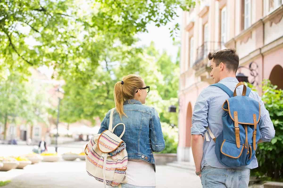 A boy on the right and a girl on the left with backpacks on walking down a sidewalk with trees on the left and a brick two-story building on the right.