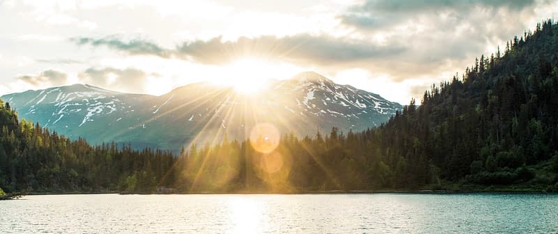 Photo of blue mountains with the sun peaking out the top of snow capped Mountains, overlooking a lake surrrounded by pine trees in Oregon, where Aldrich Wealth provides wealth, individual, and corporate retirement plan services.
