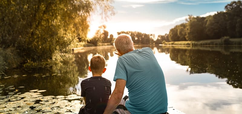 Grandfather and grandson sitting on a dock by a lake watching beautiful sunset.