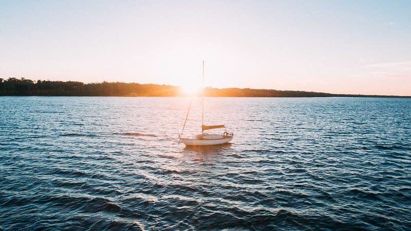Sunset overlooking the blue tater of a lake, where there is a sailboat in the middle of the water.