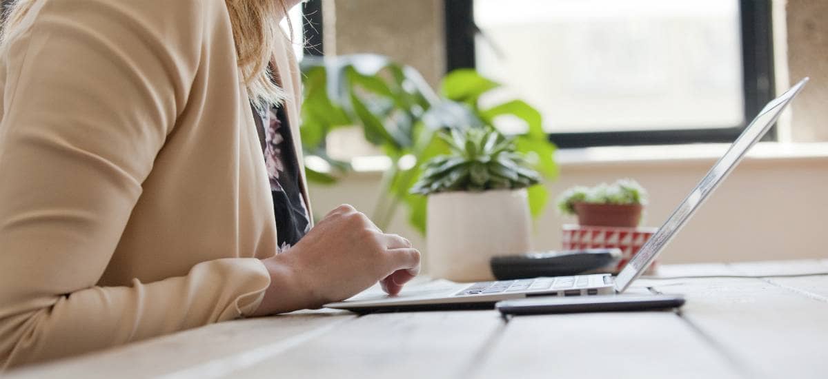 A woman with a beige jacket scrolling on her laptop, with plants in the background, in an office space. Plan fiduciaries need to be aware of the new guidance on cybersecurity threats.