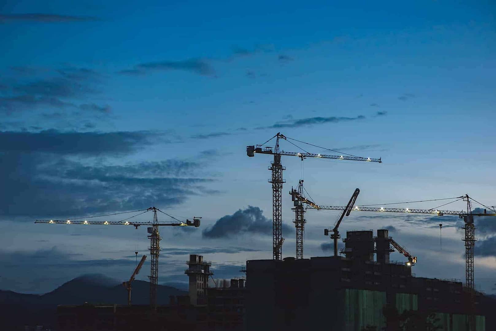 The skyline of three tall construction cranes with dark skies before daybreak.