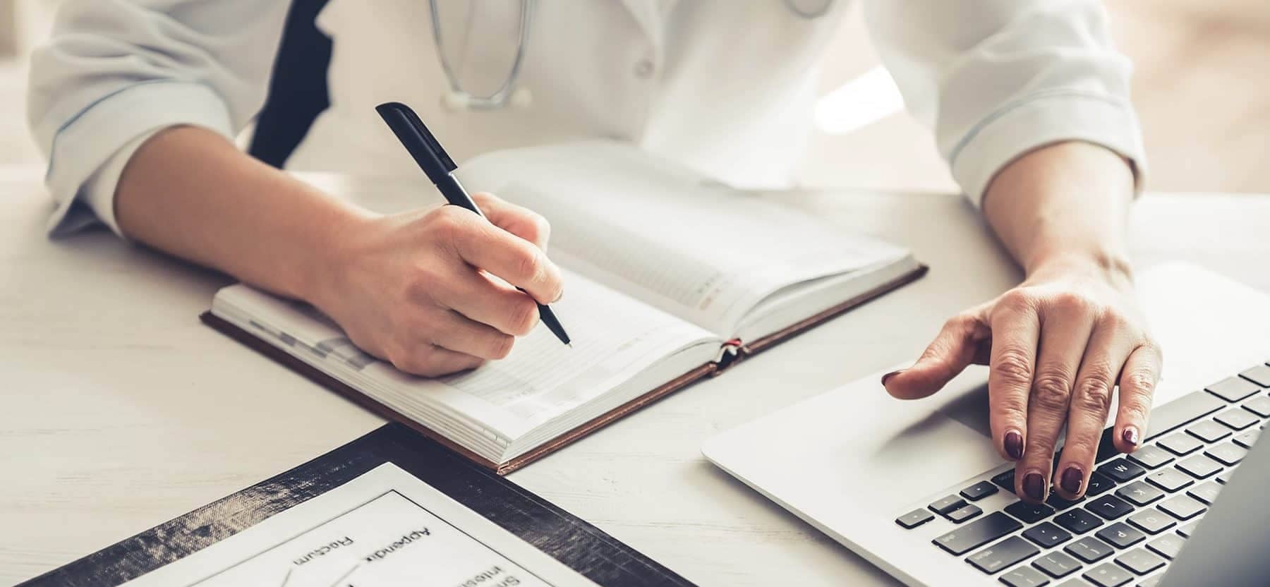 A physician on his laptop while writing on a notebook.