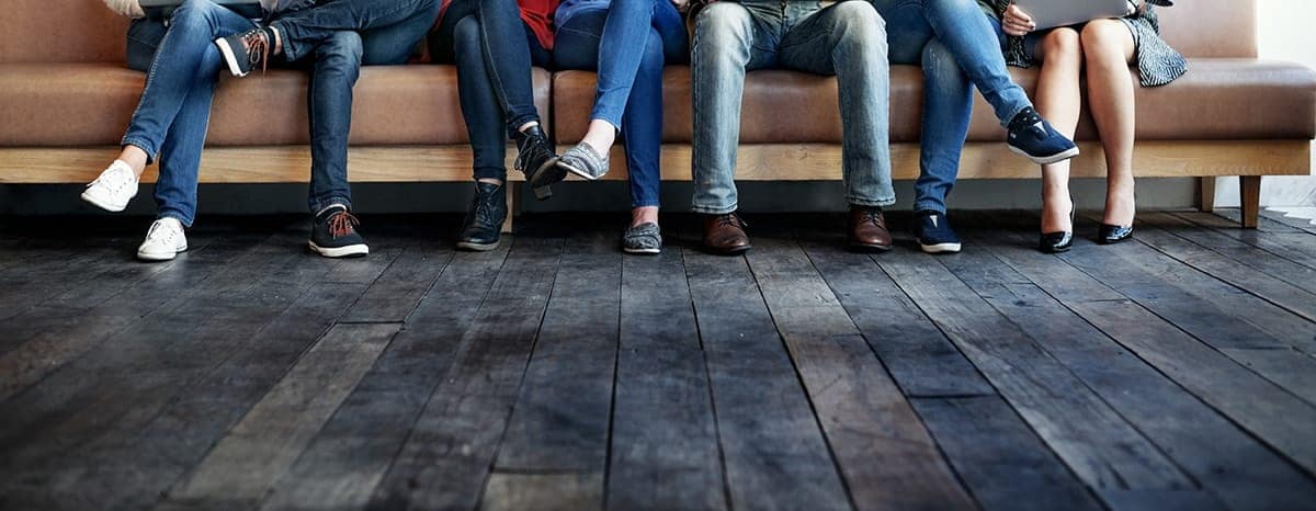 Six people's jean-covered legs, sitting on a beige couch with their feet resting on gray wooden floors.