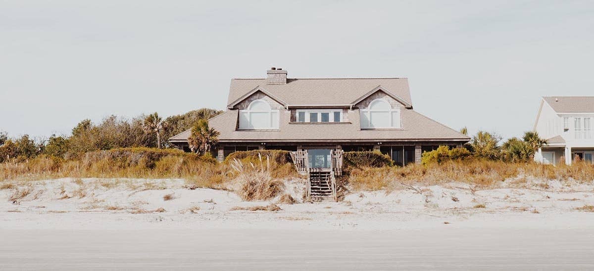 A two story beige house with steps leading down to a sandy beach shore with greenery surrounding.