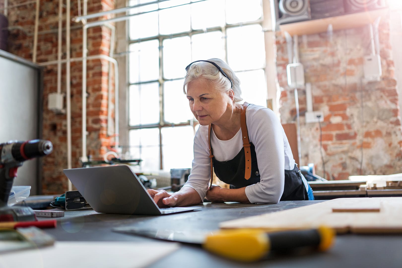 Older woman in her workshop on a laptop with a large window in the background