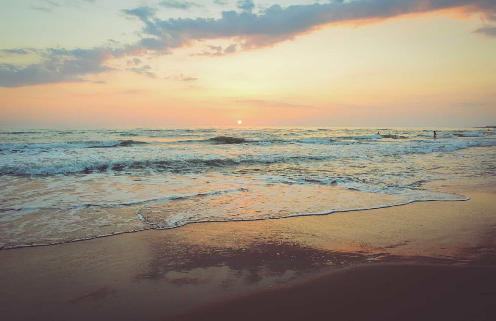 The shoreline and flowing waves of the ocean at sunset with the sun setting on the horizon with clouds in the sky.