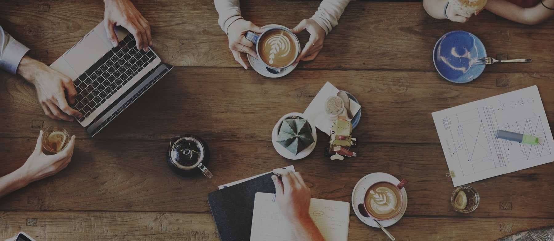 An top-down image of a wooden table and five people's hands with coffees, notebooks, and laptops around.