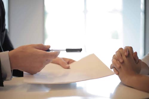 close-up image of two sets of hands across each other on a table, with one offering the other pen and paper in an M&A advisory setting. Business owners should be aware of one-on-one conversations with a prospective buyer.