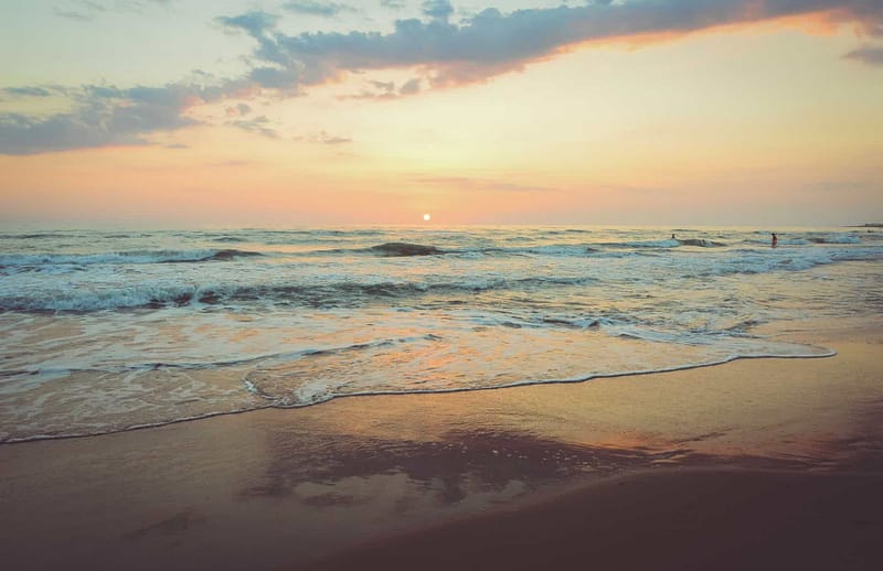 The shoreline and flowing waves of the ocean at sunset with the sun setting on the horizon with clouds in the sky.