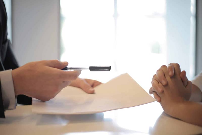 close-up image of two sets of hands across each other on a table, with one offering the other pen and paper in an M&A advisory setting. Business owners should be aware of one-on-one conversations with a prospective buyer.