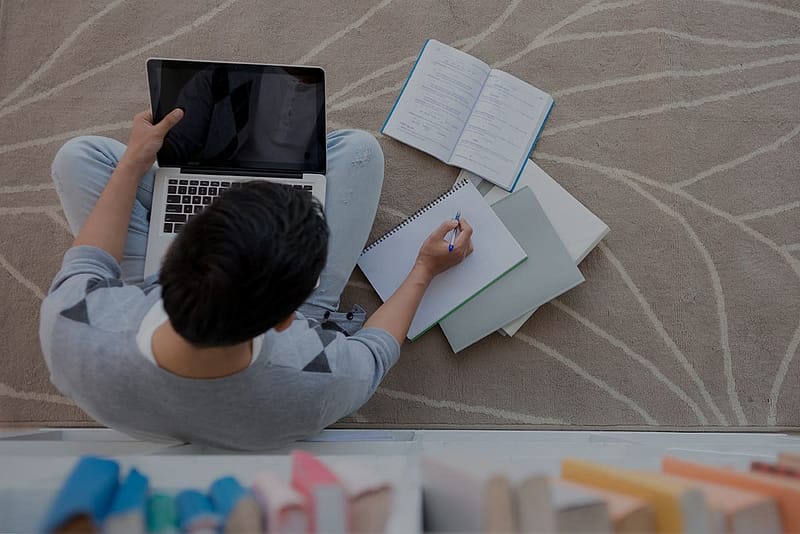A boy sitting on a beige printed carpet with a laptop in their lap and books to their right with a pencil in their hand.