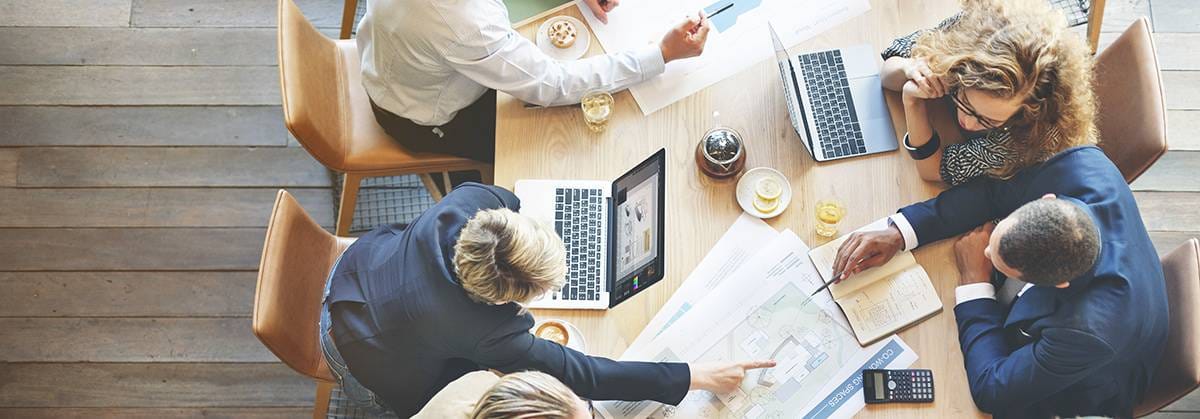 A wooden meeting table with people pointing to a large drafted document on the table and with coffees and laptops.