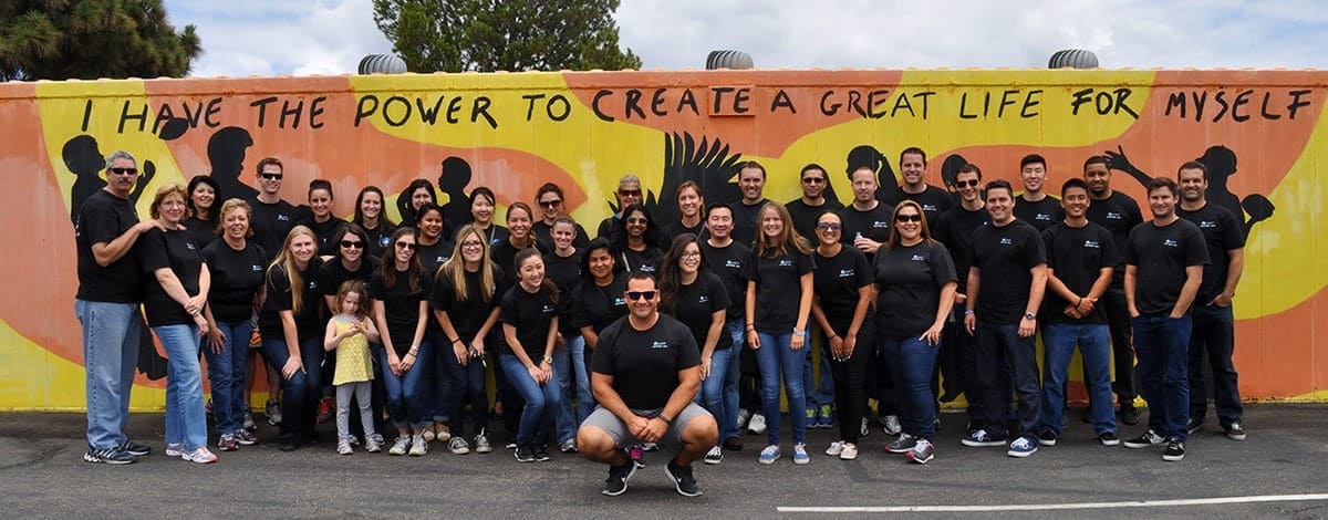 A group photo of some of Aldrich's employees in black Aldrich t-shirts in front of a orange and yellow mural.