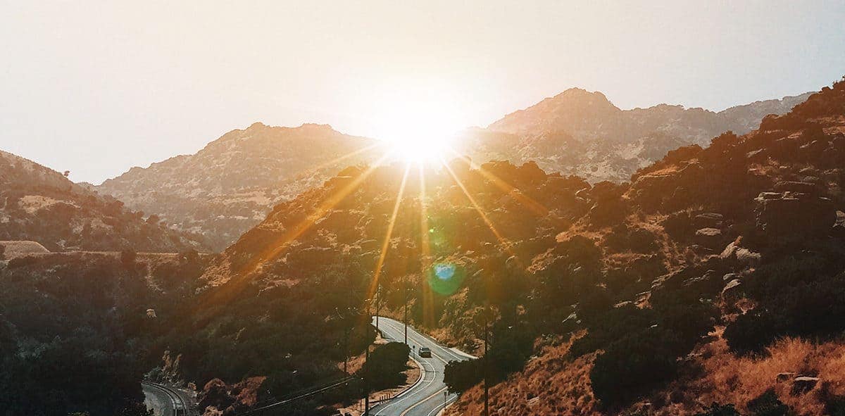 The sun peaking out of the middle of the mountains with a river in the center.