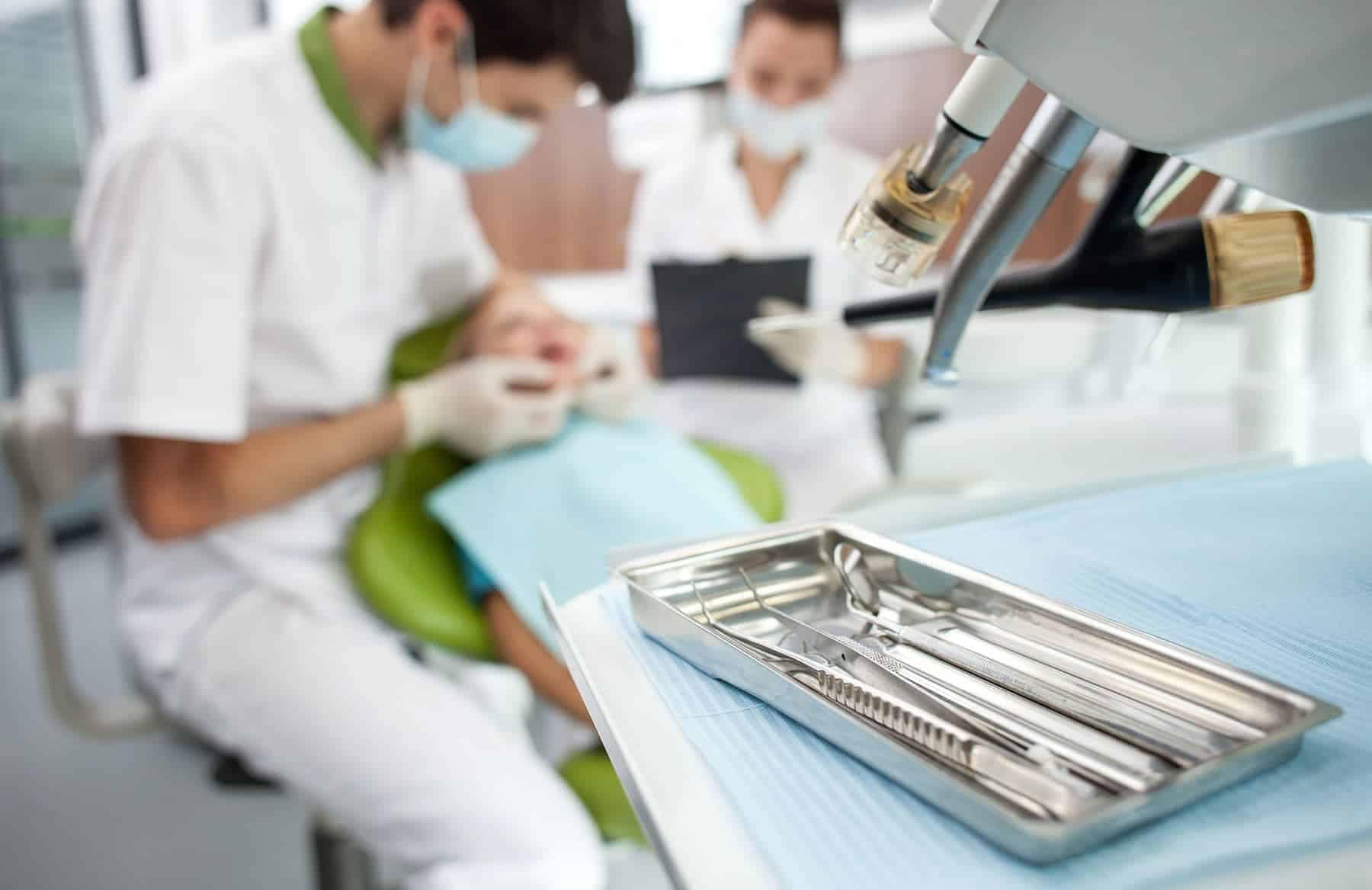 Professional young dental doctor is treating patient, with dental tools, and a woman writing on a clipboard.
