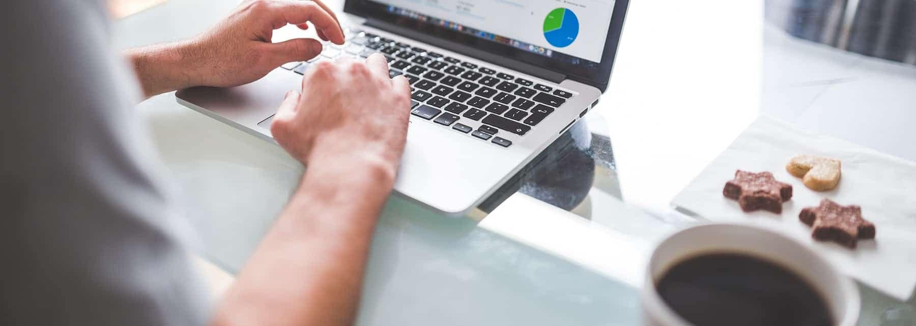 Person on a laptop on a white table with cookies and coffee to their right.