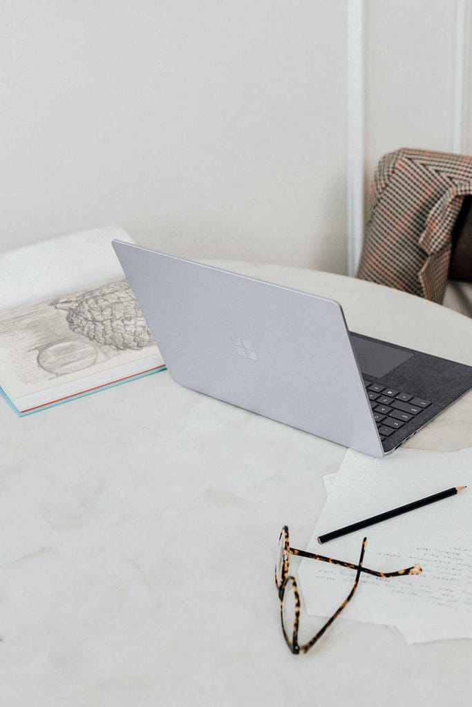 A laptop, a notebook, a pencil, and glasses on a desk with a jacket covered chair.