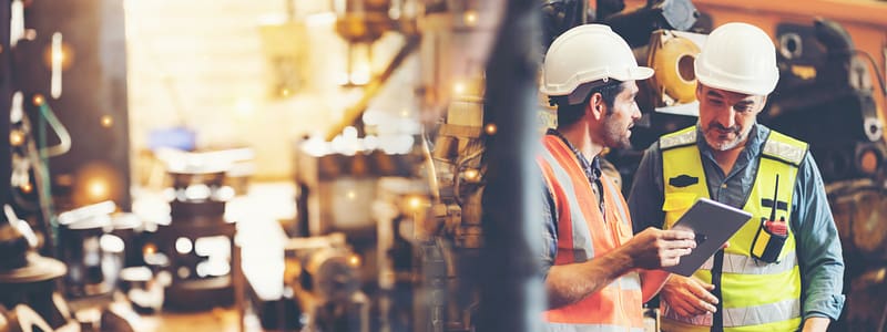 Two manufacturing workers on the shop floor with hard hats and safety vests on looking at an iPad.