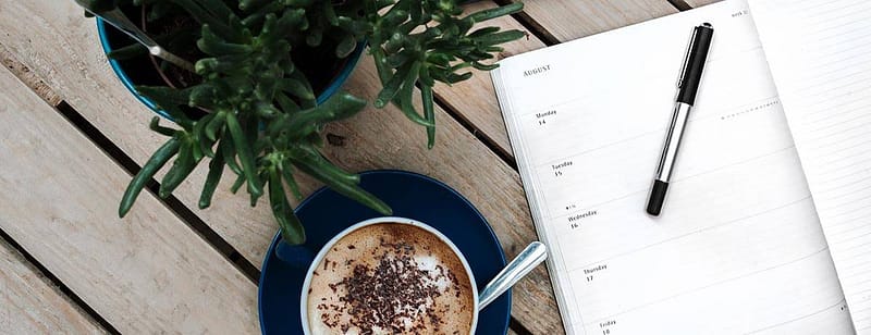 A top down view of a weekly planner book, a cup of coffee on a blue saucer with a spoon, and a plant on a wooden table.