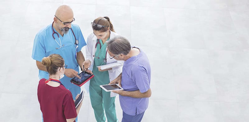 A group of a medical team in different colored scrubs discussing something while looking at their computer tablets.