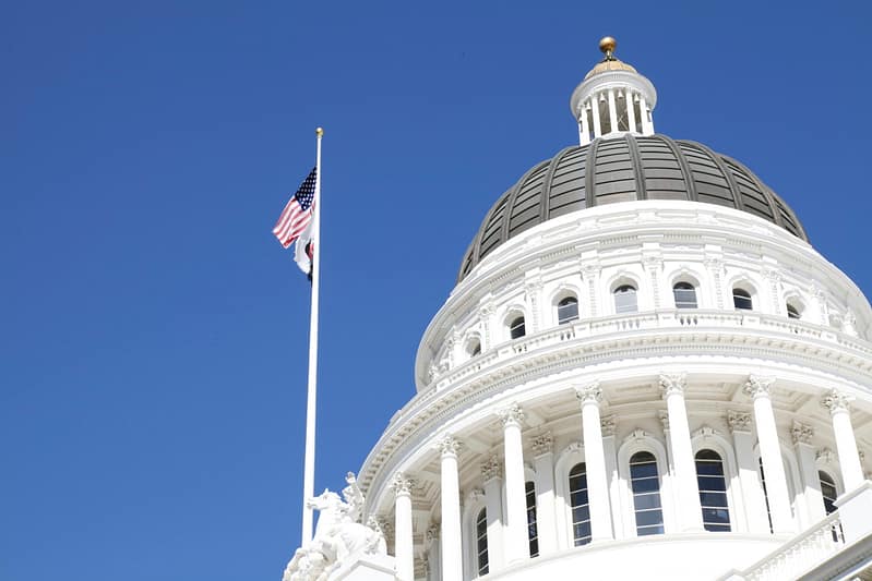 Photo of a white US government building with an American flag on a flag pole and clear blue skies in the back ground.