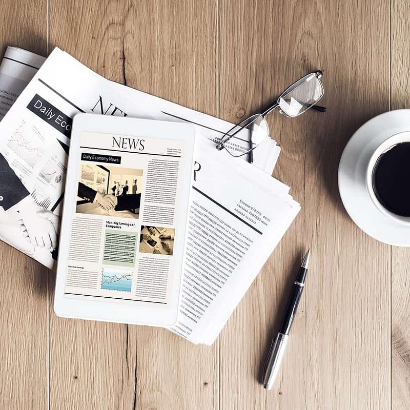 A pile of papers with images and text on a wooden table with a white cup of coffee on a saucer, a pen, and a pair of black glasses.