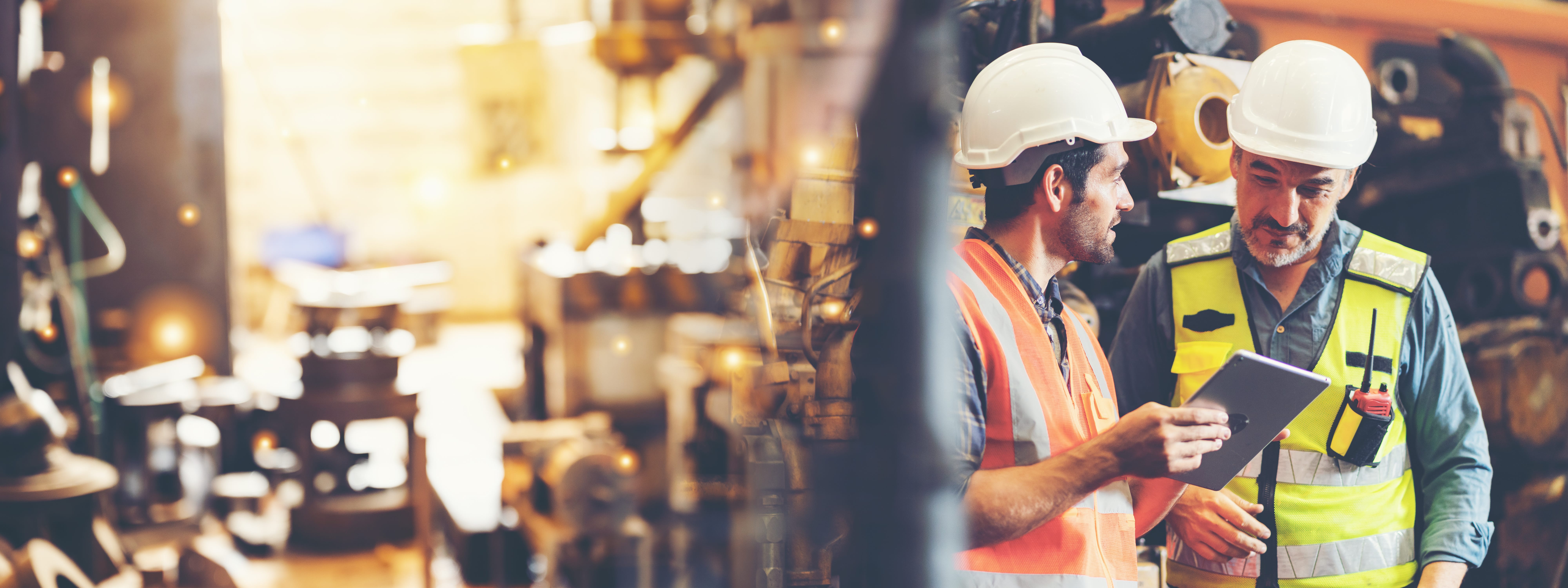 Two manufacturing workers on the shop floor with hard hats and safety vests on looking at an iPad.