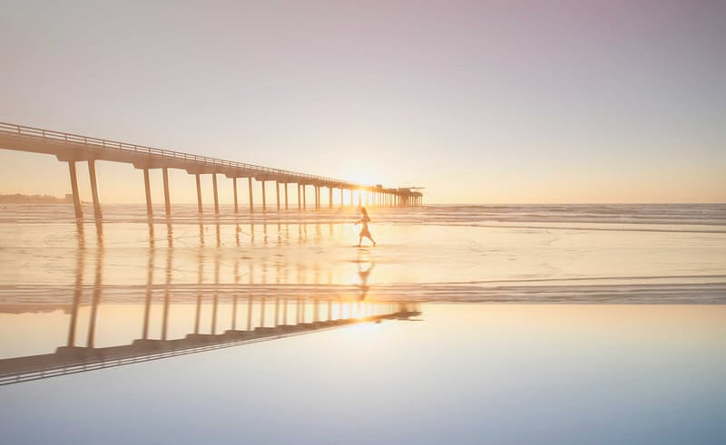 A photo of a woman walking on a San Diego beach at sunset, where the shore relects the horizon, and also where Aldrich two San Diego metro area offices.
