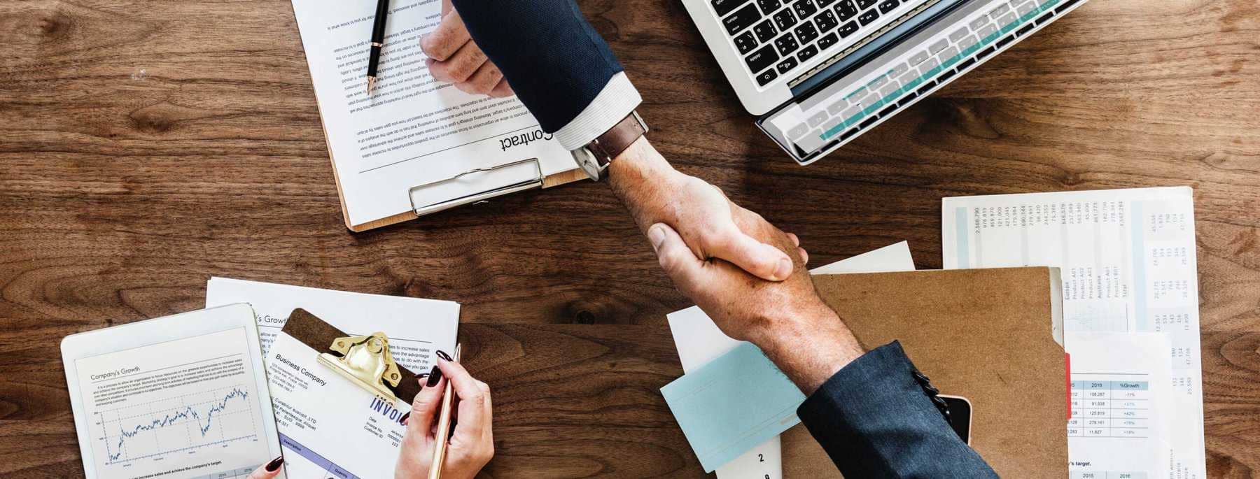 Business professionals shaking hands in office setting with paperwork and computer