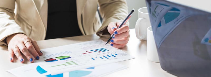 Women at office space working at a desk with laptop and pointing at a paper chart of reports, wearing tan coat and black shirt.