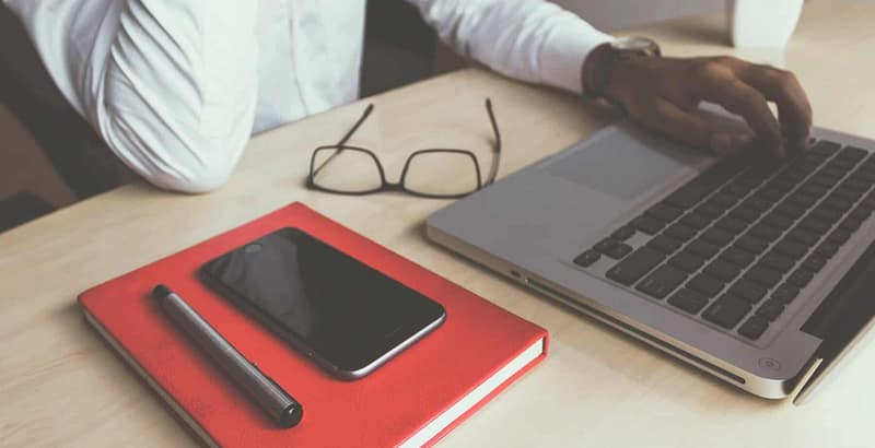 hands on a light colored wooden desk with a red notebook and macbook