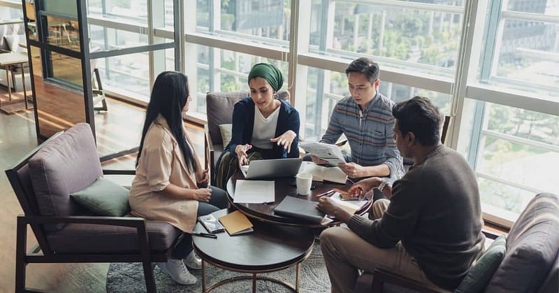 Group of collages collaborating in an office face with laptops, papers, notebooks and large windows.