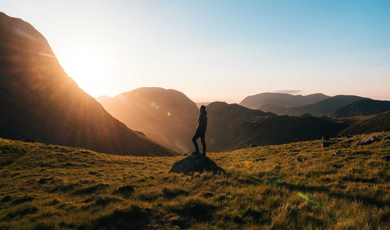 A person standing on a rock in the mountains overlooking the sunset peeking over the mountaintops.